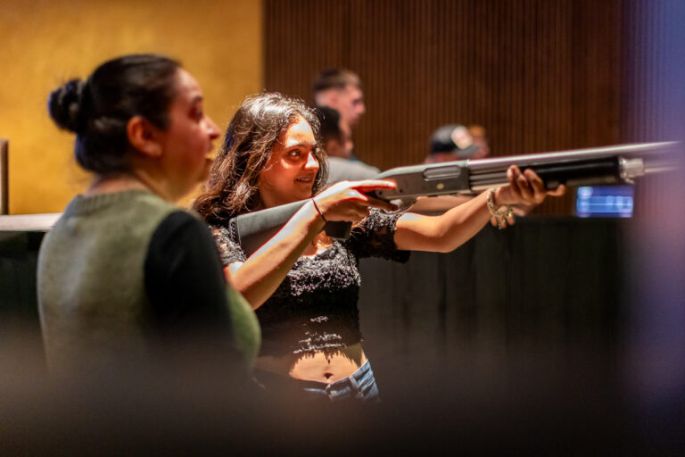 Woman in a lace black top aiming a shotgun indoors at a shooting range, with onlookers in the background