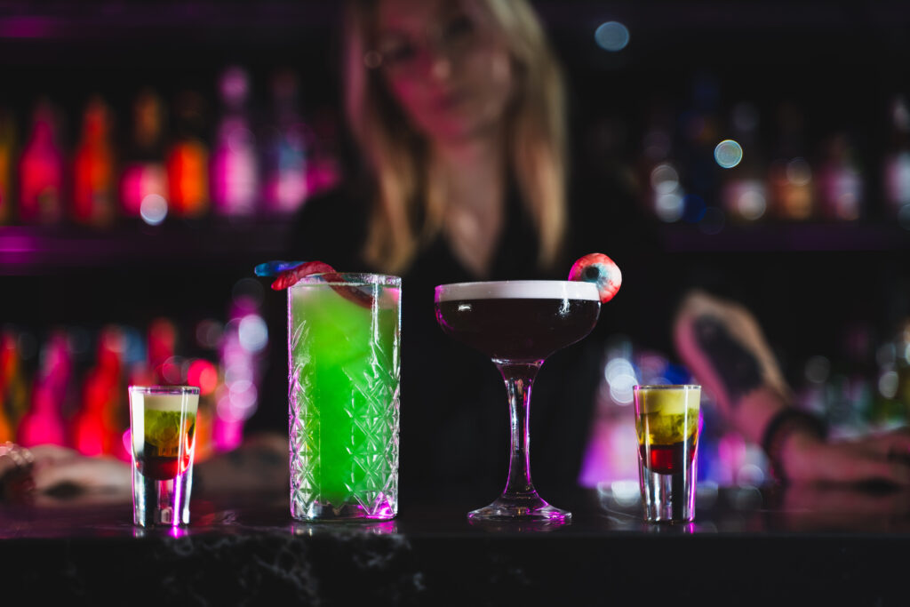 Assorted vibrant cocktails on a bar counter with a neon-lit, out-of-focus background; a tall green drink, a dark purple cocktail, and smaller shots garnished with fruit.
